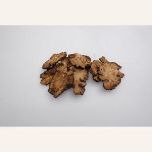 May include: A small cluster of dried, light brown root slices with irregular, jagged edges and a textured, woody surface. The slices are thin and flat, displaying a natural, fibrous appearance with varying shades of tan and dark brown throughout.