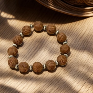 May include: A beaded bracelet featuring round, textured brown wooden beads with etched geometric patterns, alternating with small, polished silver-toned metal spacer beads. The beads are arranged in a circular loop on a light-colored wooden surface.