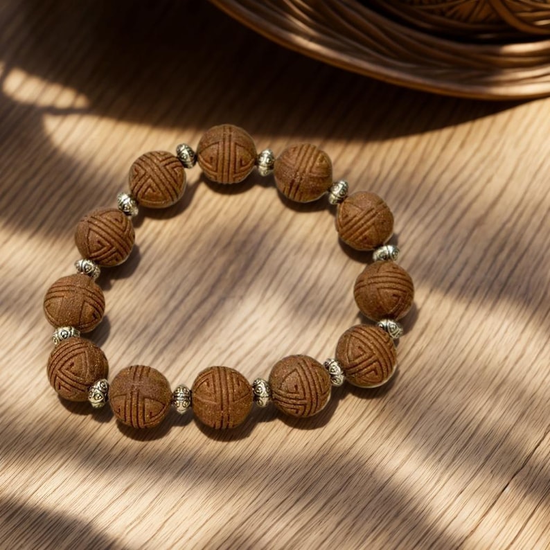May include: A beaded bracelet featuring round, textured brown wooden beads with etched geometric patterns, alternating with small, polished silver-toned metal spacer beads. The beads are arranged in a circular loop on a light-colored wooden surface.