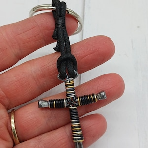 May include: A black leather keychain with a silver and gold cross made of horseshoe nails. The cross is wrapped with black and gold wire.
