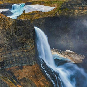 May include: A cascading waterfall flows over dark, rugged basalt cliffs into a deep canyon. Patches of white snow and ice cling to the rocky terrain, while the surrounding landscape features golden-brown grasses and mossy textures under a bright sky.