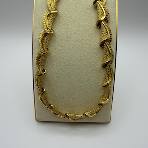 May include: A gold-tone necklace featuring a series of interconnected, curved, textured links. Each link has a distinct, repeating pattern with a matte, granular finish, creating a continuous, spiralling, ribbon-like structure around the neck.
