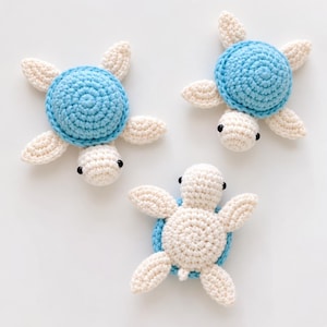 May include: Three small crocheted sea turtles with light blue shells and cream-colored heads and flippers. Each turtle features two small black bead eyes. Two turtles are positioned with their shells facing up, while one is flipped over.