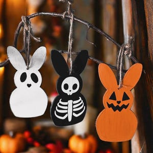 May include: Three wooden bunny ornaments hanging from a branch. The bunnies are painted in different Halloween themes: a white ghost, a black skeleton, and an orange pumpkin.