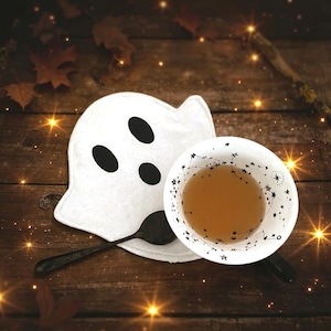 Könnte beinhalten: Ein weißer, geisterförmiger Untersetzer mit schwarzen Augen und ein schwarzer Löffel neben einer weißen Teetasse mit Tee. Die Teetasse hat ein schwarzes Sternbild-Design. Die Artikel befinden sich auf einer Holzoberfläche, Halloween-Thema.