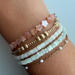 May include: A stack of five bracelets featuring a strand of peach-colored star-shaped beads, a gold-toned chain with ribbed spherical beads, two strands of square white mother-of-pearl beads, and a thin gold chain with small white round beads.