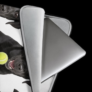 May include: A laptop sleeve featuring a black Labrador retriever holding a bright yellow tennis ball in its mouth. The sleeve is partially open, revealing a silver laptop inside. The interior of the sleeve is lined with a soft, light gray fabric.