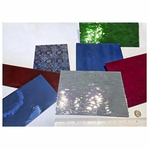 May include: Assortment of glass sheets in various colors and textures, including green, blue, gray, and red. The sheets are arranged on a white surface with a ruler and a coin for scale.