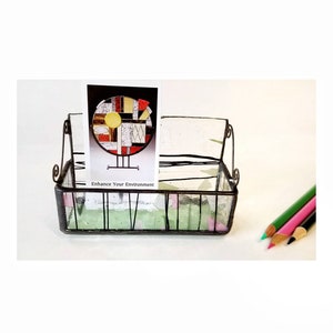 May include: A black metal wire basket with a glass base and pink and green accents. The basket is holding a card with a colorful geometric design and the text "Enhance Your Environment".
