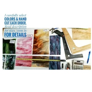 May include: Assortment of stained glass pieces in various colors and patterns, including green, pink, brown, and blue. The pieces are laid out on a white surface with measuring tape, a ruler, a calculator, and other tools.