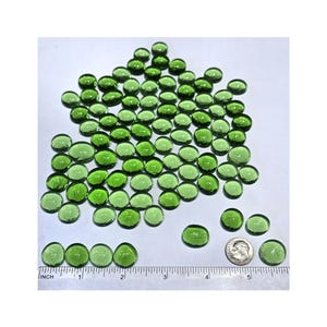 May include: A large quantity of small, oval-shaped, green glass beads. The beads are scattered on a white surface with a ruler for scale. The beads are approximately 1/2 inch in diameter.