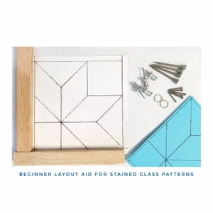 May include: A beginner's layout aid for stained glass patterns. The aid is a white sheet of paper with black lines drawn on it to create a geometric pattern. The aid is placed on a wooden frame. There are also some small metal tacks and nails on a white surface with a blue grid.