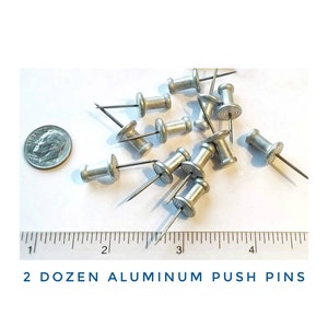 May include: Two dozen silver aluminum push pins, scattered on a white surface, next to a ruler and a US dime coin. The push pins have a round head with a small, sharp point.