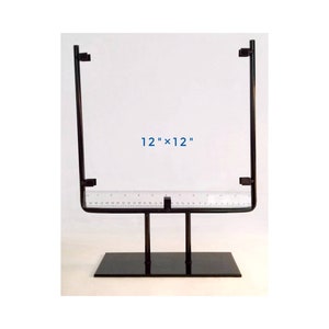 May include: A black metal display stand with a rectangular base and a square frame. The frame is 12" x 12" and includes a ruler along the bottom edge. The stand is designed to hold items for display.