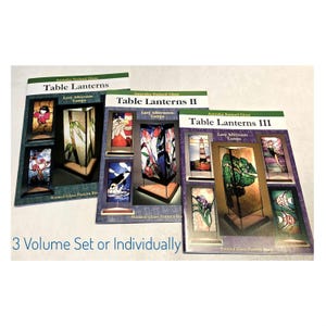 May include: Three softcover books titled Aanraku Stained Glass Table Lanterns, Table Lanterns II, and Table Lanterns III. Each cover displays various stained glass lamp designs with colorful patterns. The text 3 Volume Set or Individually is visible.