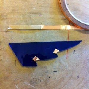May include: A blue glass piece with three small pieces of copper tape attached to it. The glass piece is on a wooden surface.