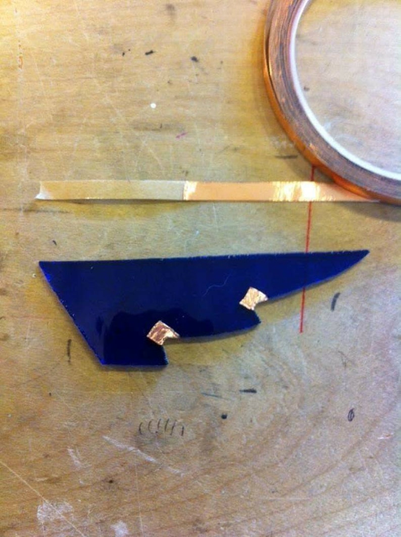 May include: A blue glass piece with three small pieces of copper tape attached to it. The glass piece is on a wooden surface.