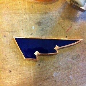 May include: A dark blue stained glass piece with a copper border. The piece is shaped like a wing and has a jagged edge.