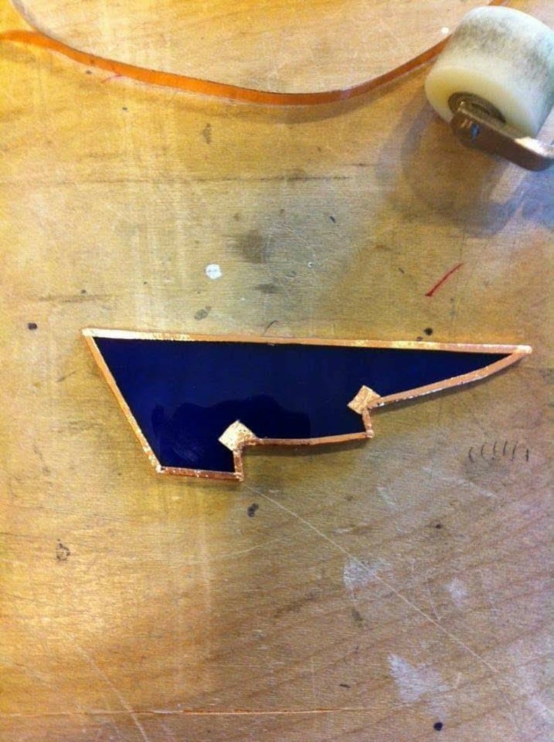 May include: A dark blue stained glass piece with a copper border. The piece is shaped like a wing and has a jagged edge.