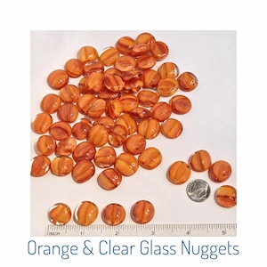 May include: A collection of orange and clear glass nuggets. The nuggets are oval-shaped with a striped pattern. A ruler and a dime are included for size reference. The text "Orange & Clear Glass Nuggets" is at the bottom.
