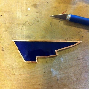 May include: A dark blue glass piece with a gold border, shaped like a wing, on a wooden surface.