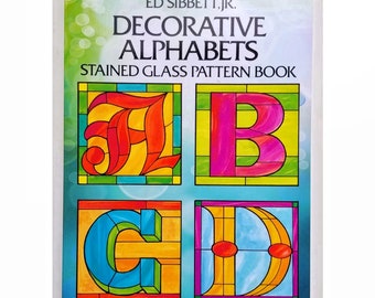May include: A book cover with the title "Decorative Alphabets" and the subtitle "Stained Glass Pattern Book" by Ed Sibbett, Jr. The cover features four colorful stained glass letters: A, B, C, and D.