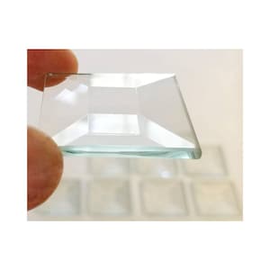 May include: A close-up of a clear glass tile with beveled edges. The tile is square and has a slightly frosted appearance. The tile is being held by a hand, and there are other tiles in the background.