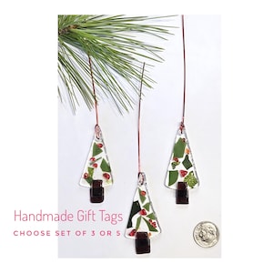 May include: Three handmade glass Christmas tree gift tags with red and green accents, suspended by copper wire. The tags have a dark brown base and are displayed with a sprig of pine. The text "Handmade Gift Tags" is visible.