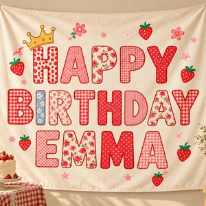May include: A cream-colored fabric wall tapestry featuring the text HAPPY BIRTHDAY EMMA in large, patterned letters. The design includes red and pink gingham, floral, and polka dot prints, accented with a yellow crown and red strawberries.