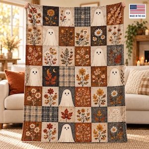 Op de afbeelding: Een rechthoekige plaid met een rasterpatroon van afwisselende vierkanten. Het ontwerp bevat witte spookillustraties, diverse herfstachtige bloemstukken, esdoornbladeren en ruitvormige stoftexturen in tinten oranje, bruin en grijs.