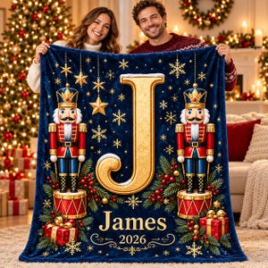 May include: A dark blue fleece blanket featuring a large gold letter J, two nutcrackers standing on red drums, pine branches, red berries, gift boxes, and gold snowflakes. The text James and 2026 are printed in gold at the bottom.