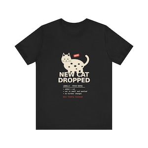 Op de afbeelding: Een zwart t-shirt met korte mouwen en een crèmekleurige afbeelding van een gevlekte kat met een rood 'NEW'-label. Onder de kat staat de tekst 'NEW CAT DROPPED', gevolgd door 'v2026.9 PATCH NOTES', drie opsommingstekens en 'devs finally listened'.