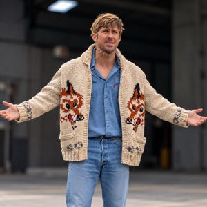 May include: A beige knit cardigan featuring a prominent fox graphic on both front panels, a shawl collar, and ribbed cuffs. The sweater is worn over a light blue button-down shirt and light blue denim jeans, creating a layered look.