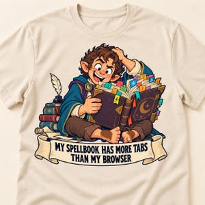 Op de afbeelding: Een crèmekleurig t-shirt met een cartoonillustratie van een fantasiefiguur met harige voeten die in een groot spreukenboek met tabbladen leest. Onder het figuur staat op een banier de tekst: MY SPELLBOOK HAS MORE TABS THAN MY BROWSER.