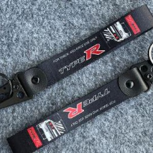 Pode incluir: Dois porta-chaves de tecido preto com o texto TYPE R em vermelho e branco, juntamente com a frase FOR TRACK AND RACE USE ONLY. Cada porta-chaves inclui um clipe de metal preto, um anel circular e uma ilustração gráfica da traseira de um carro.