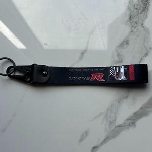 May include: A black fabric keychain strap featuring the text "TYPE R" in red and white, along with the phrase "FOR TRACK AND RACE USE ONLY" in white. The strap includes a black metal lobster clasp and a circular split ring attachment.