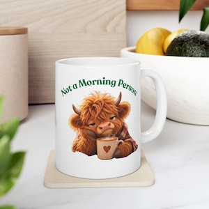 Peut inclure: Un mug en céramique blanche orné d'une illustration de vache Highland brune et poilue à l'air somnolent, tenant une petite tasse beige avec un cœur. Le texte "Not a Morning Person" est imprimé en vert au-dessus de l'illustration.