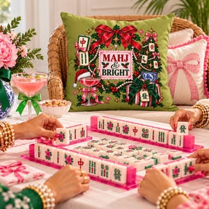 Può includere: Un cuscino quadrato verde oliva con una grafica festiva natalizia che include un fiocco rosso, un albero di Natale, un bicchiere da cocktail con un cappello da Babbo Natale e la scritta MAHJ & BRIGHT, circondato da agrifoglio e perle.