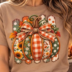 Può includere: Una t-shirt a maniche corte color marrone chiaro con una grande grafica di una zucca decorata con un fiocco a quadri. I segmenti della zucca mostrano vari motivi, tra cui girasoli, margherite bianche, quadretti rossi e bianchi e pois.