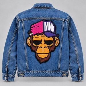 May include: A blue denim jacket featuring a large embroidered patch on the back. The patch depicts a monkey wearing sunglasses and a split-colored pink and purple baseball cap with the white text MNK printed on the front of the cap.