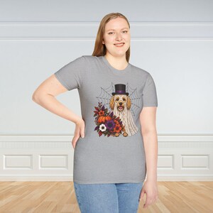May include: A heather grey short-sleeved t-shirt featuring a graphic of a golden-coloured dog wearing a white ghost sheet and a black top hat. The dog is positioned in front of a spider web and surrounded by a colourful arrangement of autumn flowers and a pumpkin.