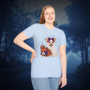 May include: A light blue short-sleeved t-shirt featuring a graphic of a dog wearing a white ghost sheet and a purple bow. The dog is positioned in front of a spider web and is surrounded by a colourful arrangement of autumn flowers and a pumpkin.