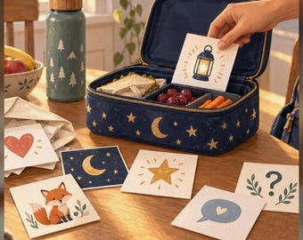 Könnte beinhalten: Eine marineblaue Lunchbox mit goldenen Sternen und einer Mondsichel, gefüllt mit einem Sandwich, Weintrauben und Karotten. Daneben steht eine türkisfarbene Trinkflasche mit weißen Bäumen und quadratische Karten mit verschiedenen Motiven.