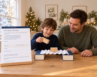 Könnte beinhalten: Ein weißes Anleitungsblatt mit der Überschrift Build a paper snow bridge steht auf einem Holzständer. Eine gefaltete weiße Papierbrücke überspannt zwei dunkelblaue Bücher, auf denen zwei kleine Holzklötze liegen.
