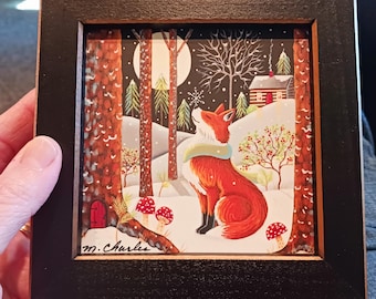 May include: A square framed print featuring a red fox wearing a light blue scarf in a snowy forest. The scene includes a full moon, pine trees, a small cabin, red mushrooms, and a tiny red door at the base of a tree. The artist signature M. Charles is visible.