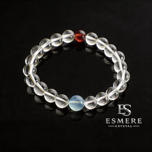 May include: A beaded bracelet featuring clear, spherical crystal beads arranged in a circular pattern. The design includes one distinct opaque light blue bead and one translucent reddish-orange bead, with the text ESMERE CRYSTAL visible below.