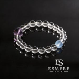 May include: A circular beaded bracelet composed of clear, translucent spherical beads. The bracelet features one distinct purple bead and one distinct light blue bead interspersed among the clear beads. The text ES ESMERE CRYSTAL is visible.