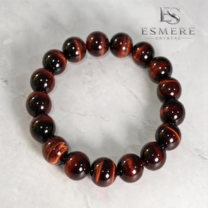 May include: A circular bracelet composed of polished, spherical red tiger's eye beads. Each bead displays a deep reddish-brown hue with characteristic chatoyant bands that reflect light. The bracelet is arranged in a ring shape on a light surface.
