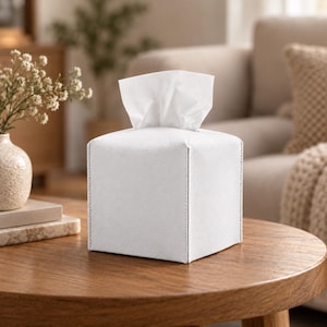 Puede incluir: Una funda cuadrada para caja de pañuelos de color blanco con un acabado texturizado similar al cuero y costuras visibles en los bordes. Un pañuelo facial blanco asoma por la abertura superior, extendiéndose hacia arriba desde el centro.