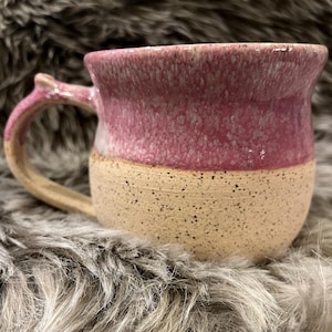 Peut inclure: Une tasse en céramique présentant un design bicolore avec une base crème mouchetée et une partie supérieure rose texturée et marbrée. La tasse possède un bord large et évasé ainsi qu'une anse incurvée assortie à la base de couleur crème.